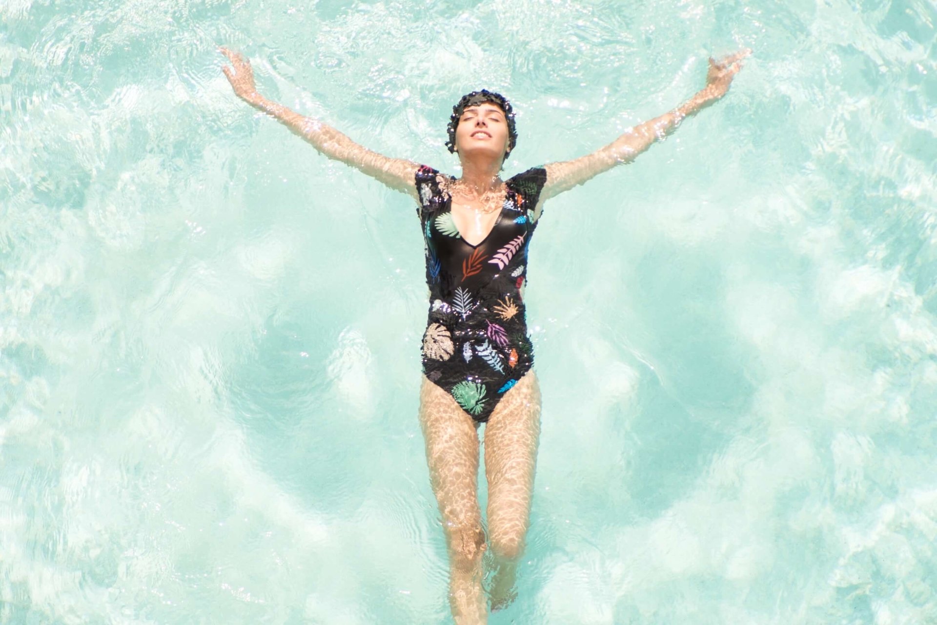 woman in one-piece bathing suit and matching swimcap floats on her back in light blue water