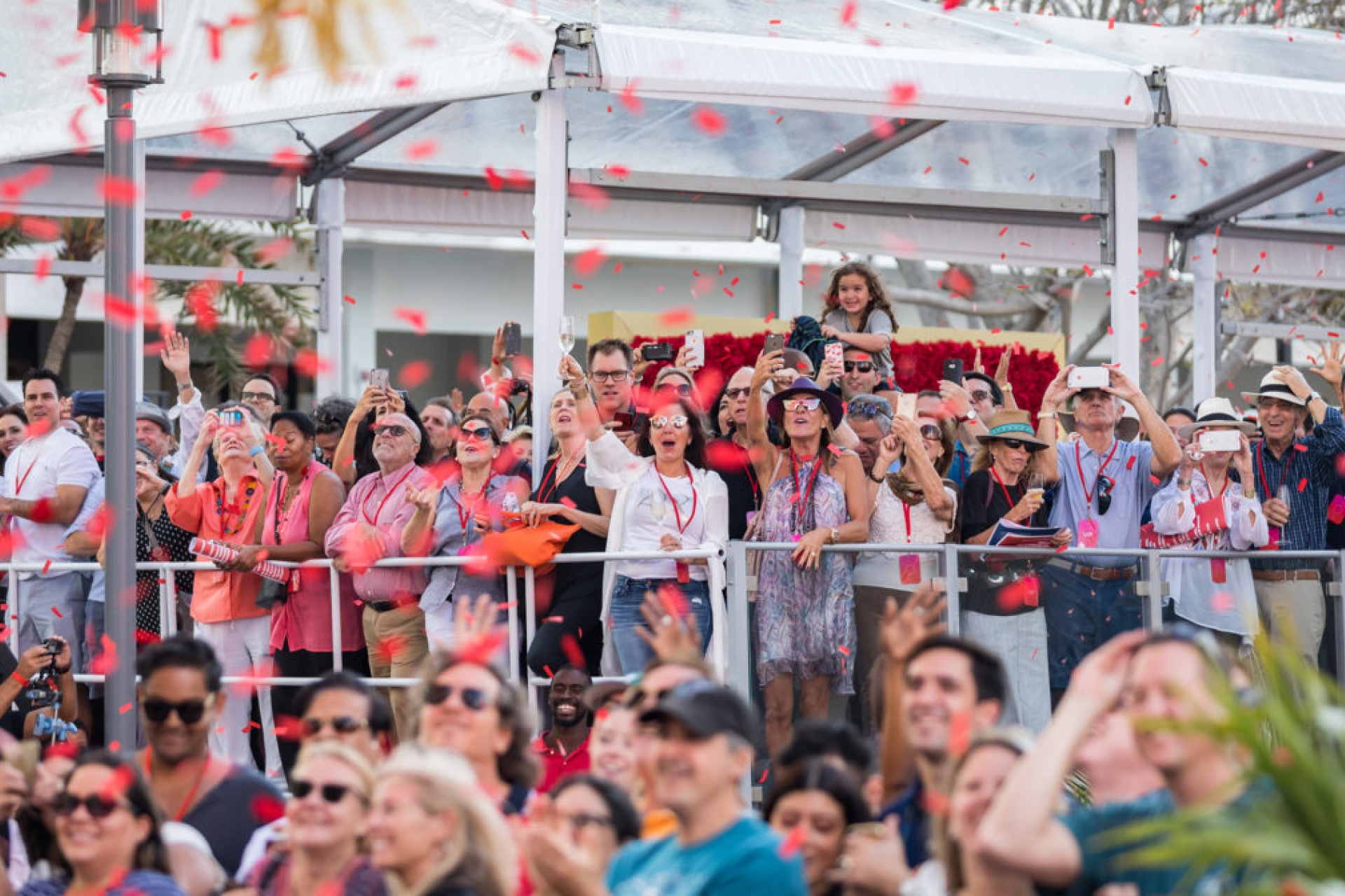 crowd stands along fence while cheering and waving with red confetti in the air