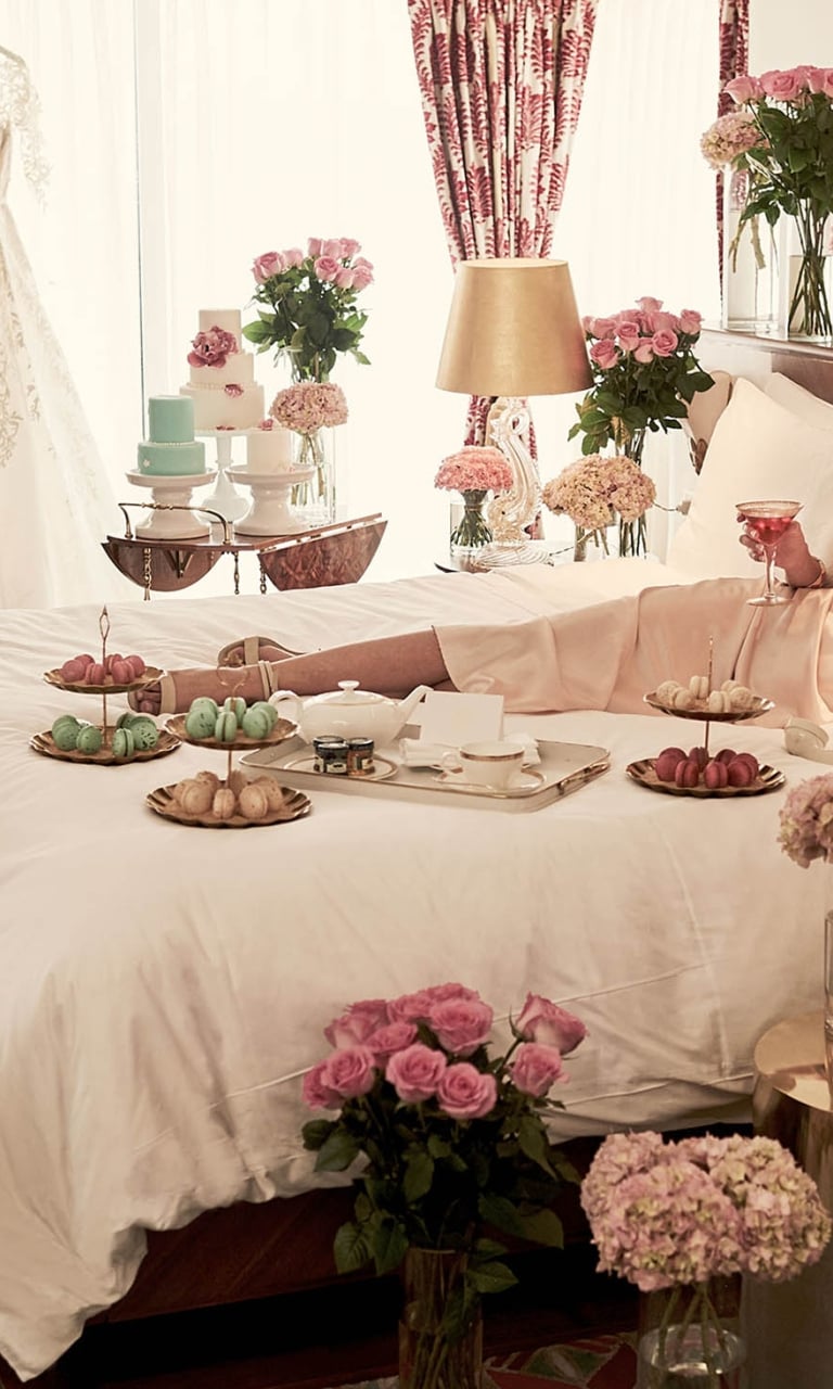 A luxurious bedroom with a large bed, decorated with pink flowers, white linens, and a selection of cakes and pastries on tiered trays.