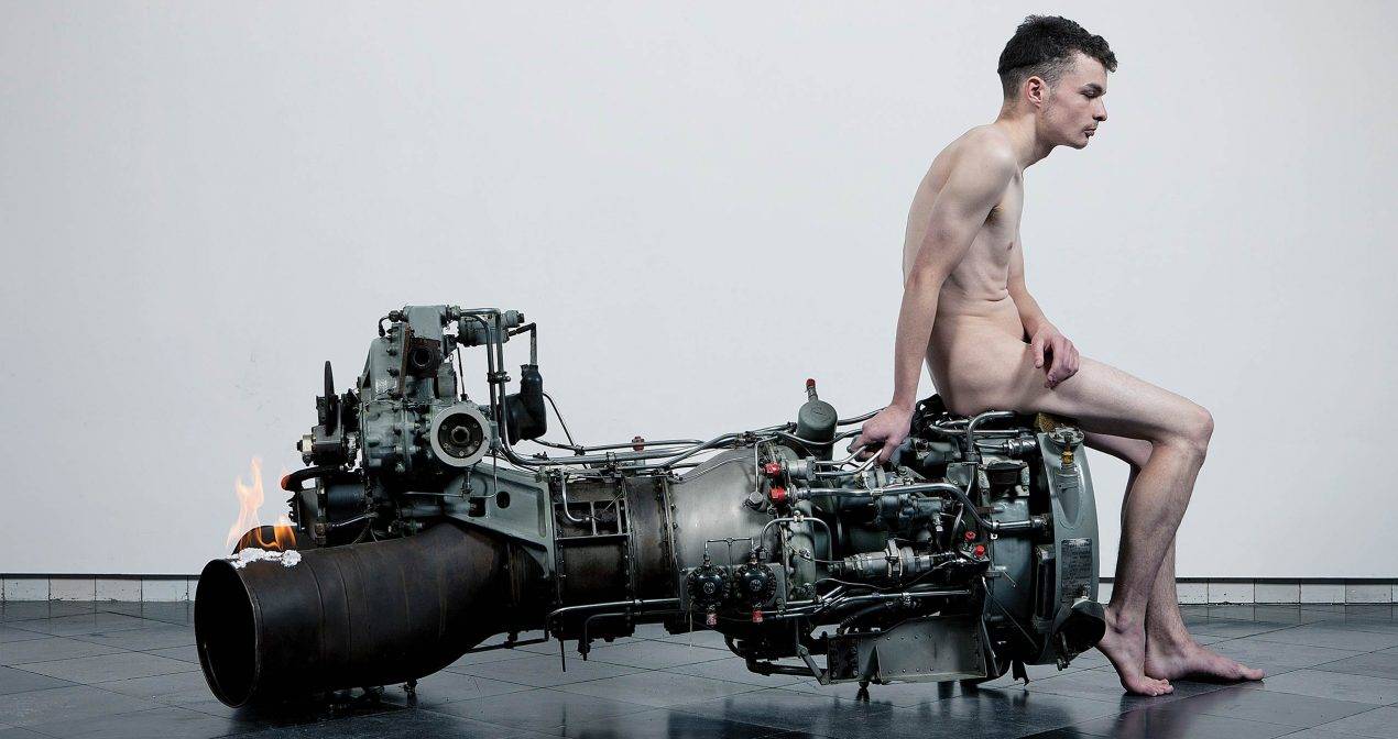 naked white man sits on the right edge of a large engine-like metal object