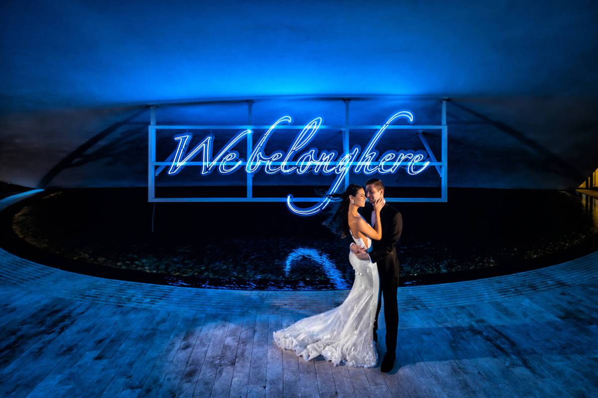 bride and groom stand in front of blue neon sign that reads "We belong here"