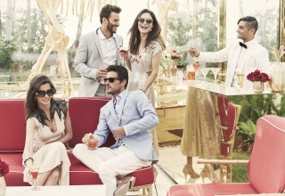 a couple sits on red bench while another stands behind them as bartender stirs cocktail on mirrored bar