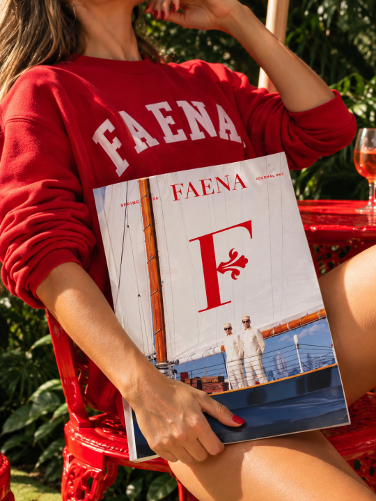 Woman wearing a red Faena sweatshirt holding a Faena Journal magazine while seated outdoors in a tropical garden.