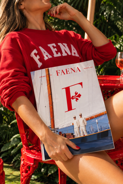 Woman wearing a red Faena sweatshirt holding a Faena Journal magazine while seated outdoors in a tropical garden.