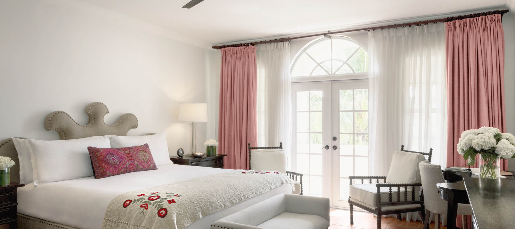 A bright and airy hotel room with a large bed, two chairs, and a table in front of a door with pink curtains.