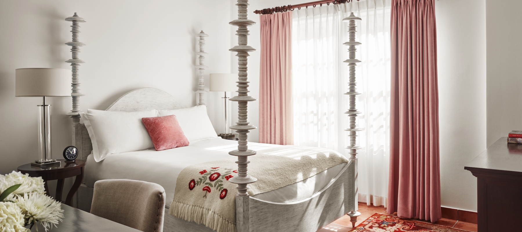 A bright and inviting hotel room with a four-poster bed, white bedding, and pink accents.