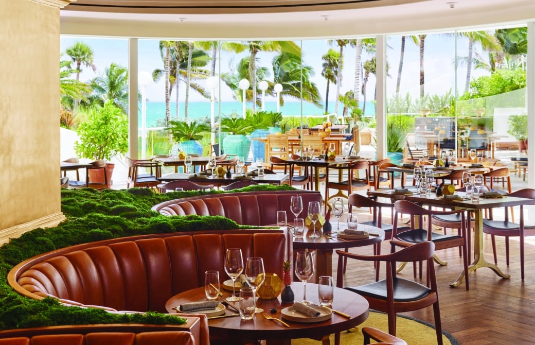 A dining area with a curved leather banquette, round tables set with wine glasses, and a large window overlooking palm trees and the ocean.