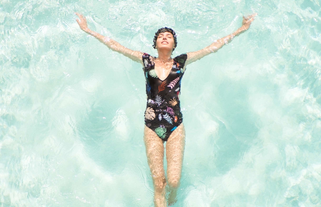 woman in one-piece bathing suit and matching swimcap floats on her back in light blue water