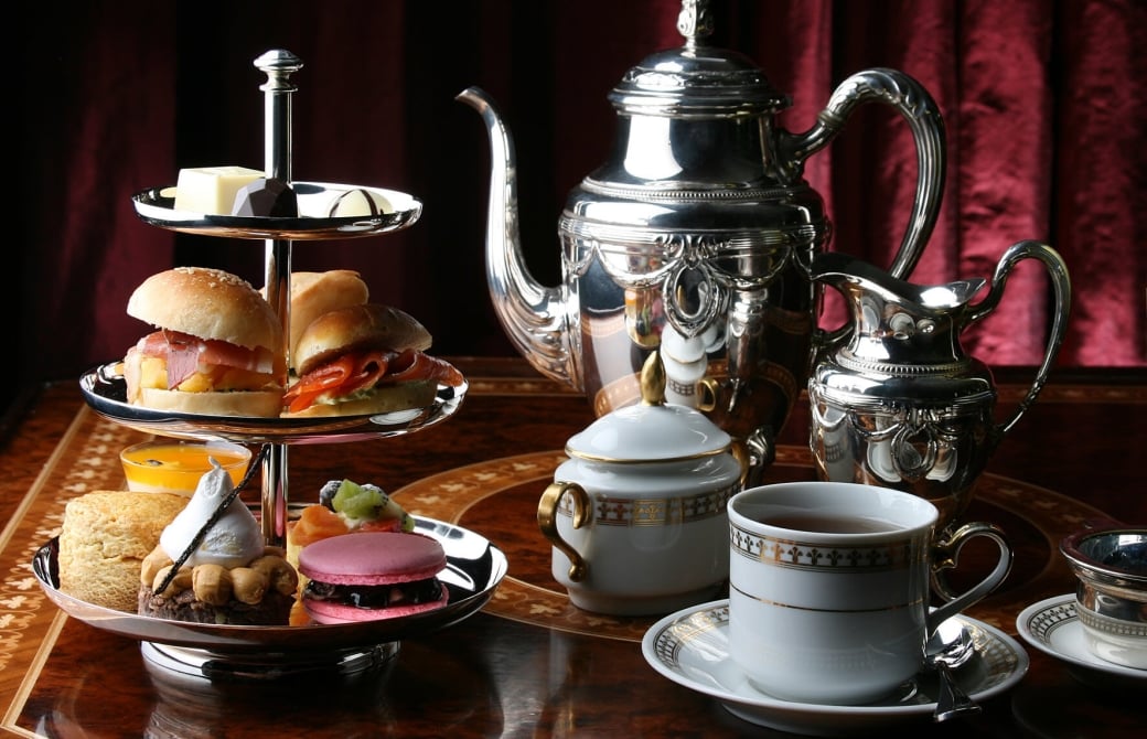 silver teapot and milk jug sit on table beside two tea cups and a multi-tiered tray of sandwiches and desserts