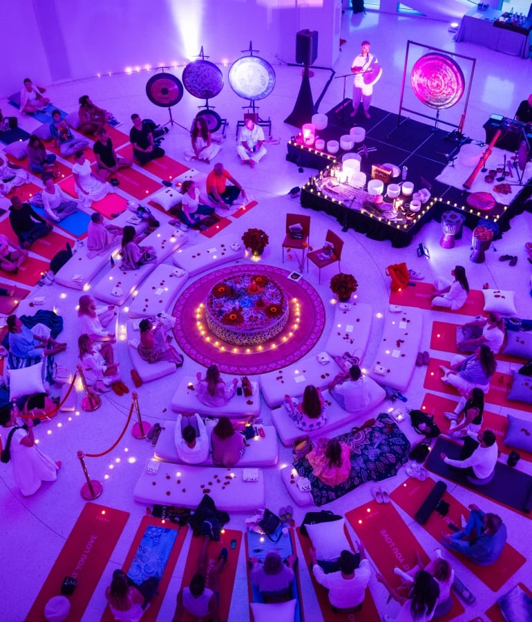 A group of people are sitting in a circle on yoga mats and cushions in a large room with purple lighting, surrounded by gongs and candles.