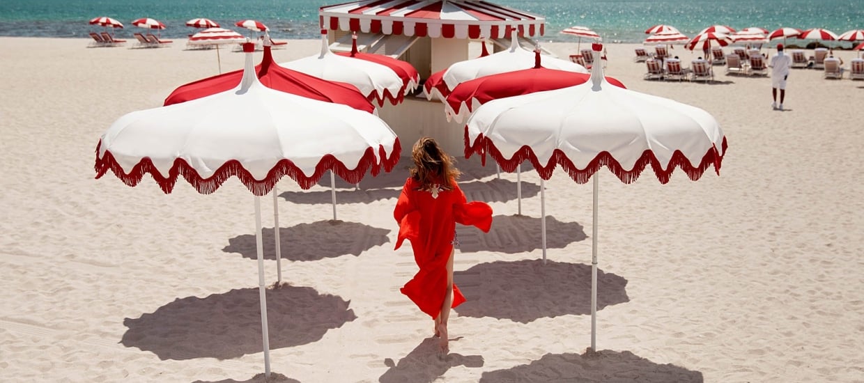A woman in a red dress walking on a beach with red and white umbrellas and ocean in the background.