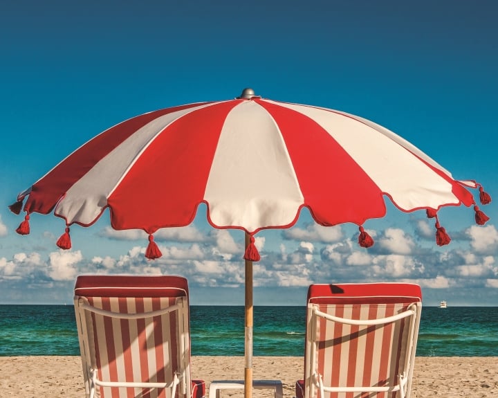 A red and white striped umbrella and two matching beach chairs on a sandy beach with an ocean view.