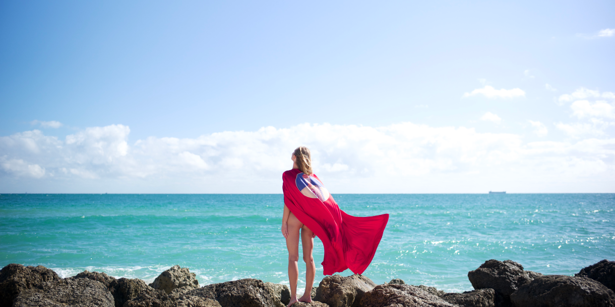 A woman stands on rocks at the beach, looking out at the ocean.