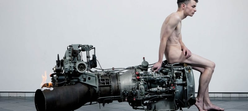 naked white man sits on the right edge of a large engine-like metal object
