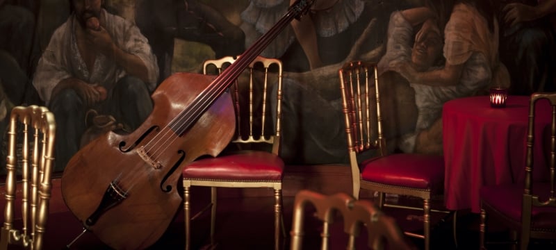 cello lays against red and gold chairs in front of wall with mural of people