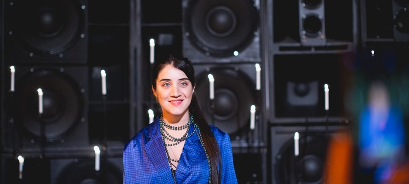 Vivian Caccuri wearing blue top stands in front of speakers