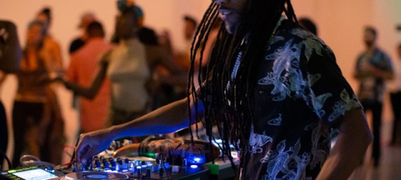 DJ Jigüe performs at DJ booth wearing hat and sunglasses