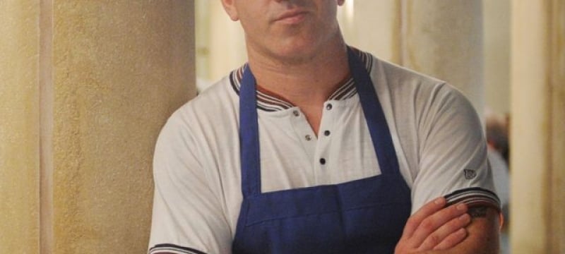 man with short hair leans on wall with arms crossed wearing chef's apron