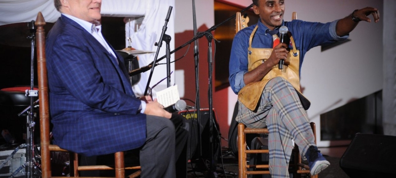 two men sit on stage, one in a suit and the other in a chef's apron, while the man in the apron speaks into a microphone
