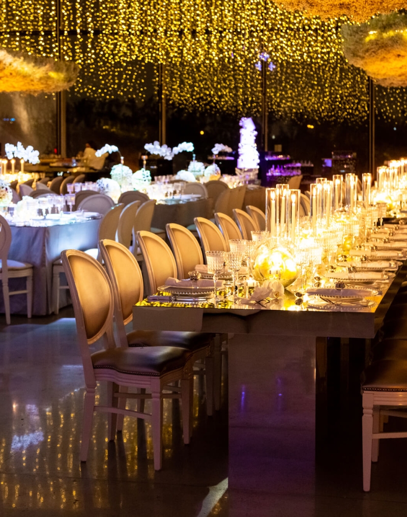 A long, elegantly set dining table with white chairs, candles, and string lights in a formal setting.