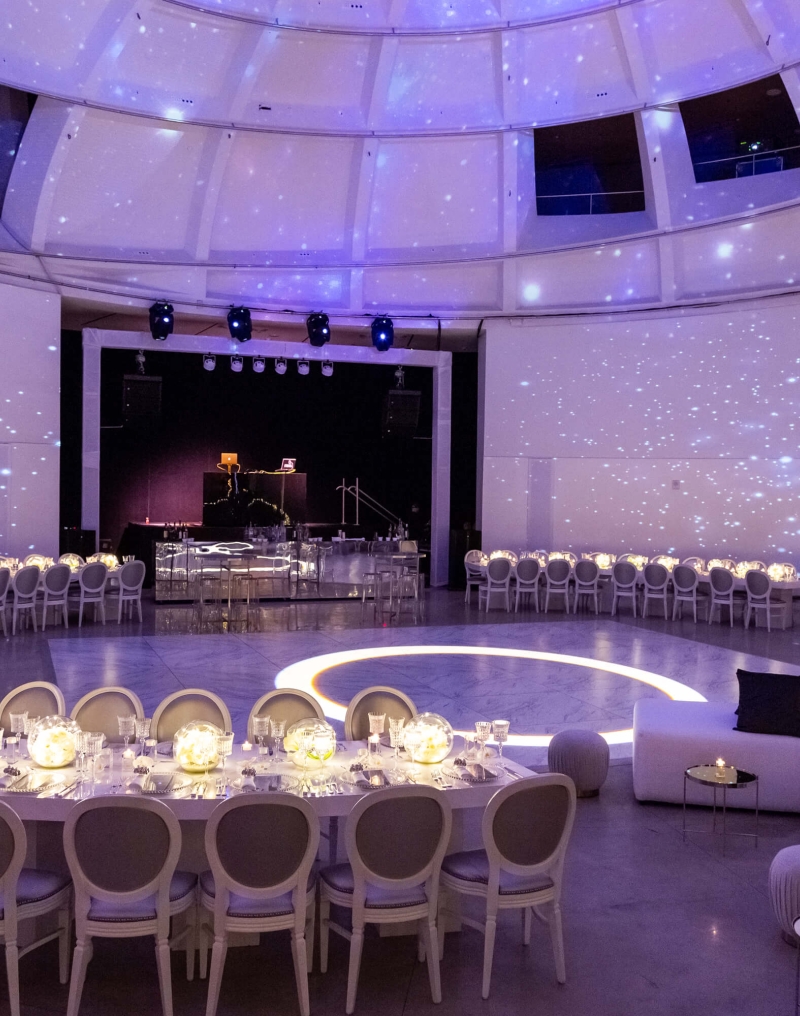 A large, circular dining room with a starry night light effect on the ceiling and walls, set with tables and chairs for a special event.