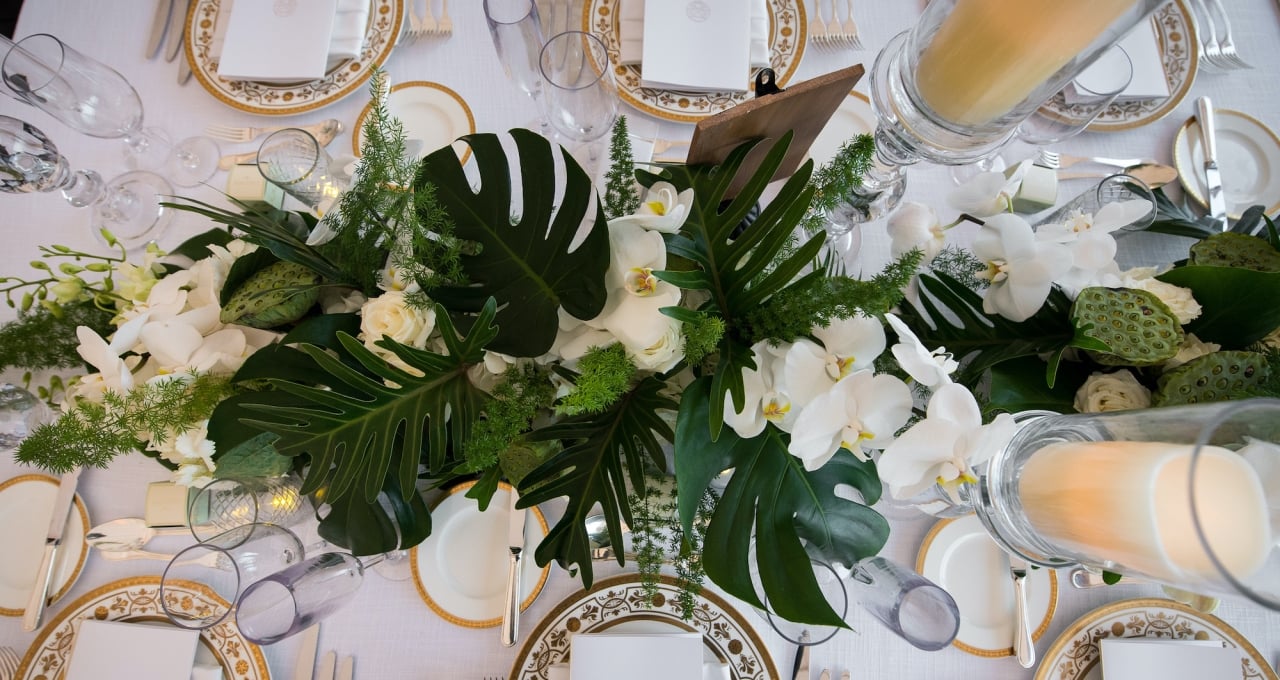 A dining table set with white flowers, green leaves, candles, plates, glasses, and silverware.