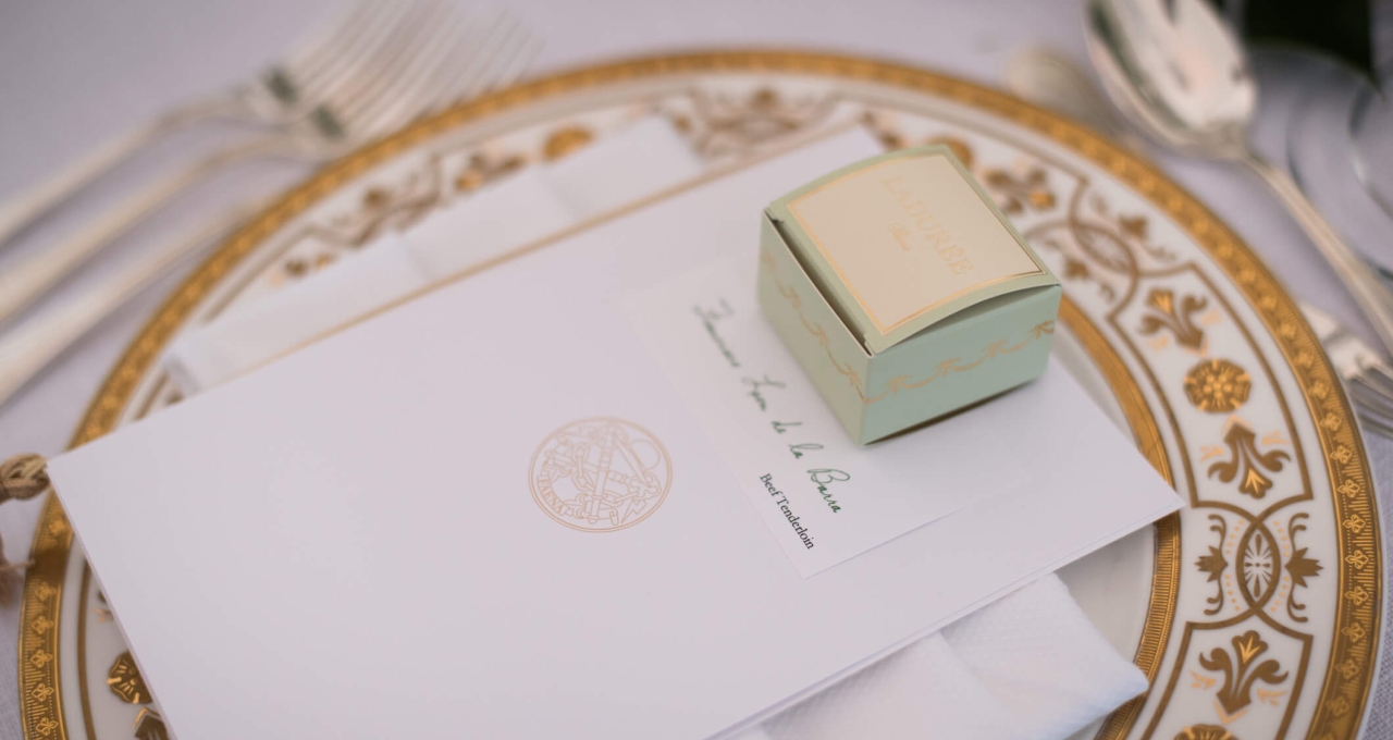 A formal table setting with a white tablecloth, gold-rimmed plate, and fine dining utensils, featuring an envelope and a small box.